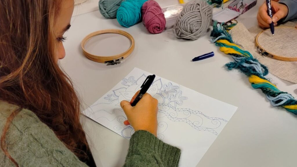 Ragazza che disegna il motivo di un ricamo creativo su carta, seduta a un tavolo con fili colorati e telai da ricamo.